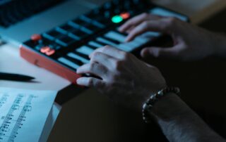 Hände spielen auf einem kleinen MIDI-Keyboard, während Notenblätter und ein Laptop im Hintergrund sichtbar sind.