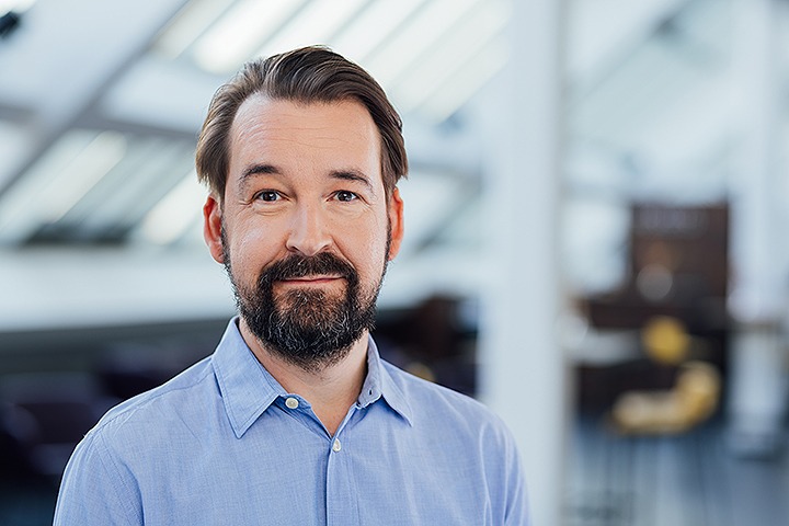Christian Rost, weißer mittelalter Mann in hellblauem Hemd mit braunem Haar und Bart lächelt leicht in die Kamera