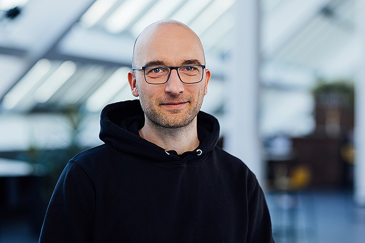 Weißer Mann mit dunklem Kapuzenpulli, Glatze und Brille lächelt in die Kamera