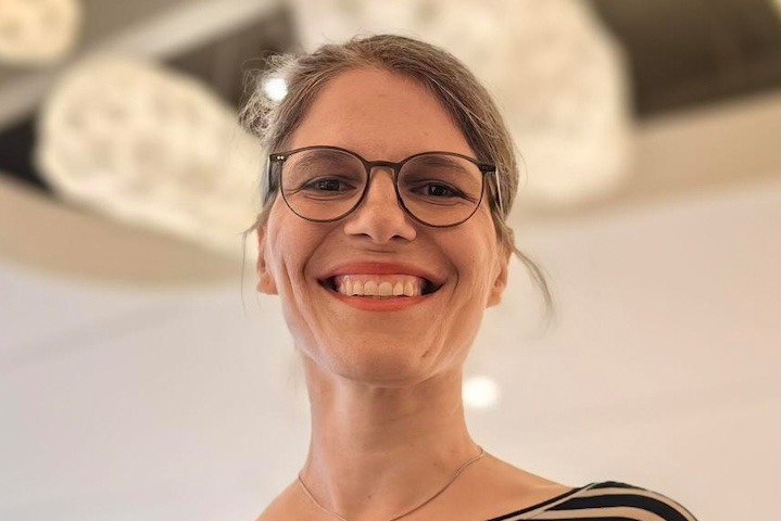 Weiße Frau mit zusammengebundenem Haar und Brille grinst etwas von oben in die Kamera
