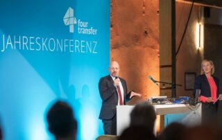 Eine Bühne mit blauem Hintergrund und der Schrift four transfer Jahreskonferenz, davor steht ein Mann im Anzug und einem Mikrofon in der Hand, daneben eine blonde Frau im Sakko.