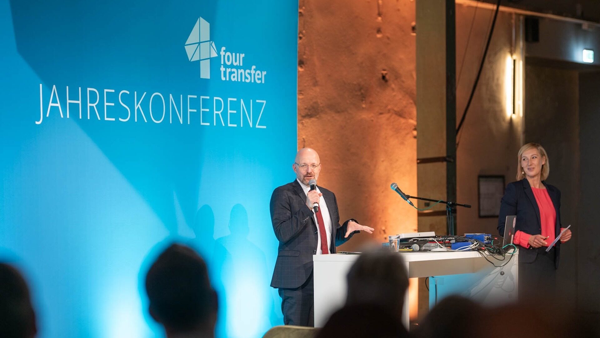 Eine Bühne mit blauem Hintergrund und der Schrift four transfer Jahreskonferenz, davor steht ein Mann im Anzug und einem Mikrofon in der Hand, daneben eine blonde Frau im Sakko.
