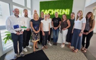 Viele weiße Personen stehen vor einem grünen Schild mit der Leuchtschrift Sachsen,