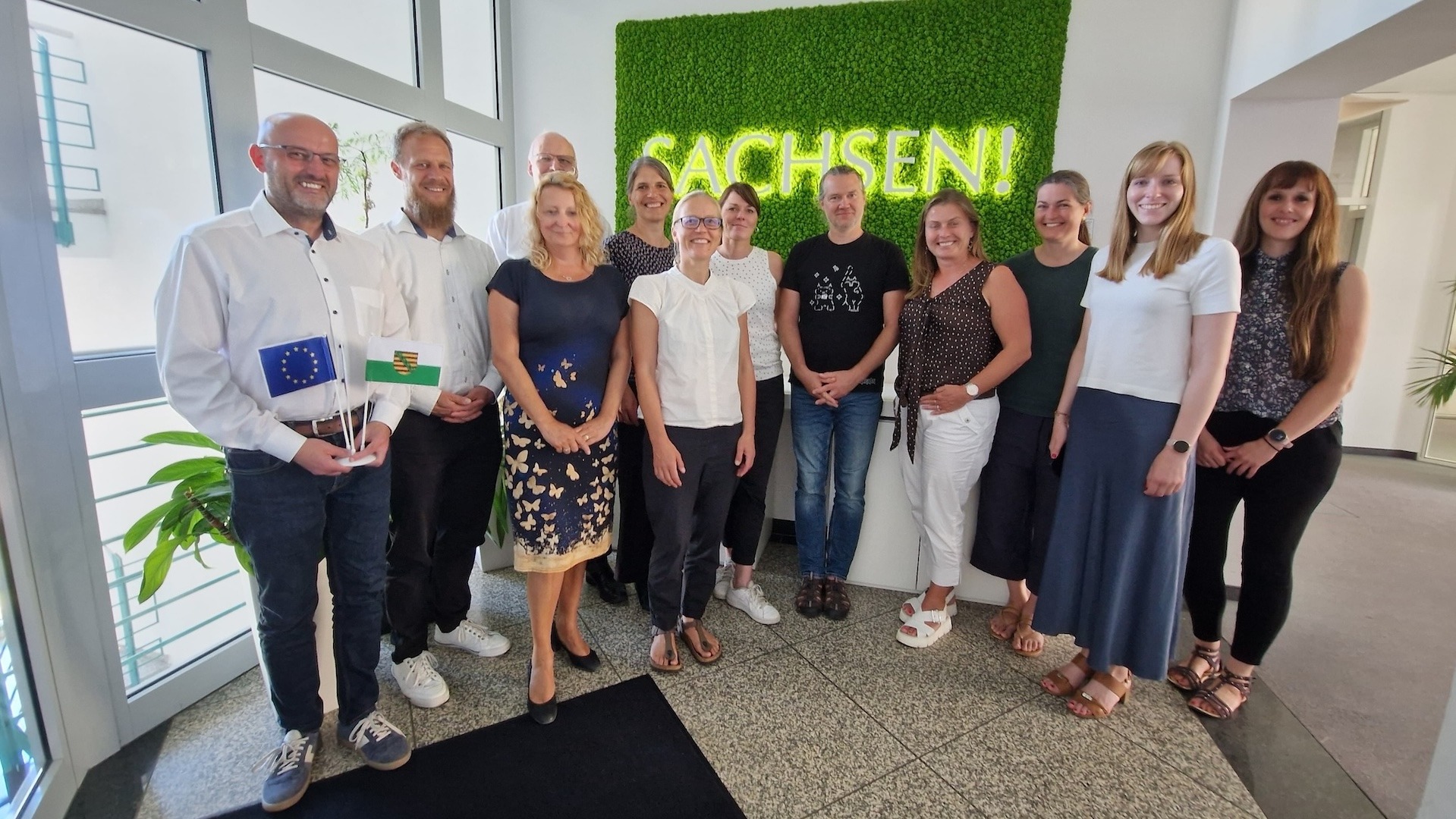 Viele weiße Personen stehen vor einem grünen Schild mit der Leuchtschrift Sachsen,